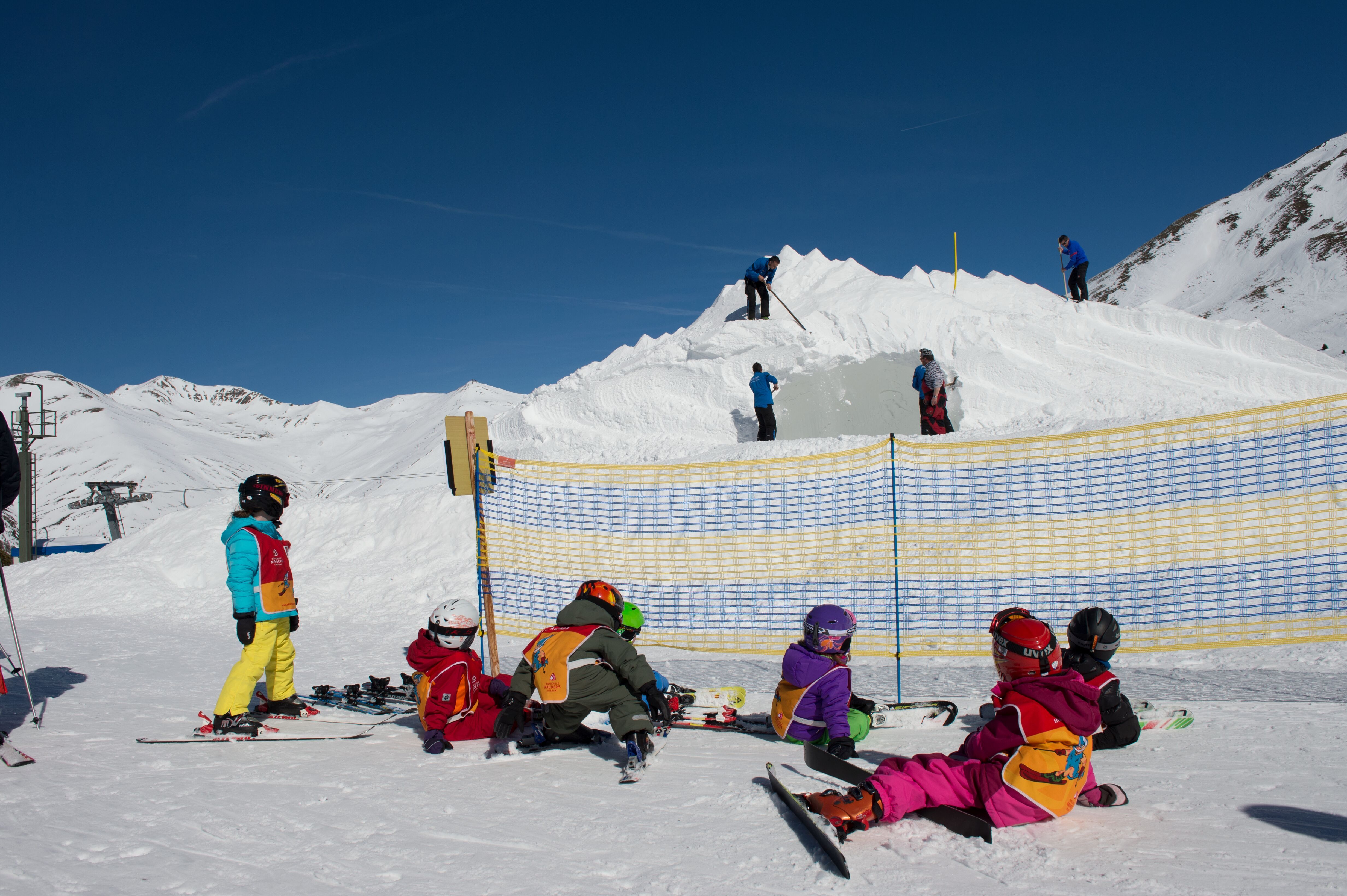 Kinder machen Pause auf dem Boden im Skigebiet, im Hintergrund bearbeiten Männer mit Schaufeln einen Schneehaufen 