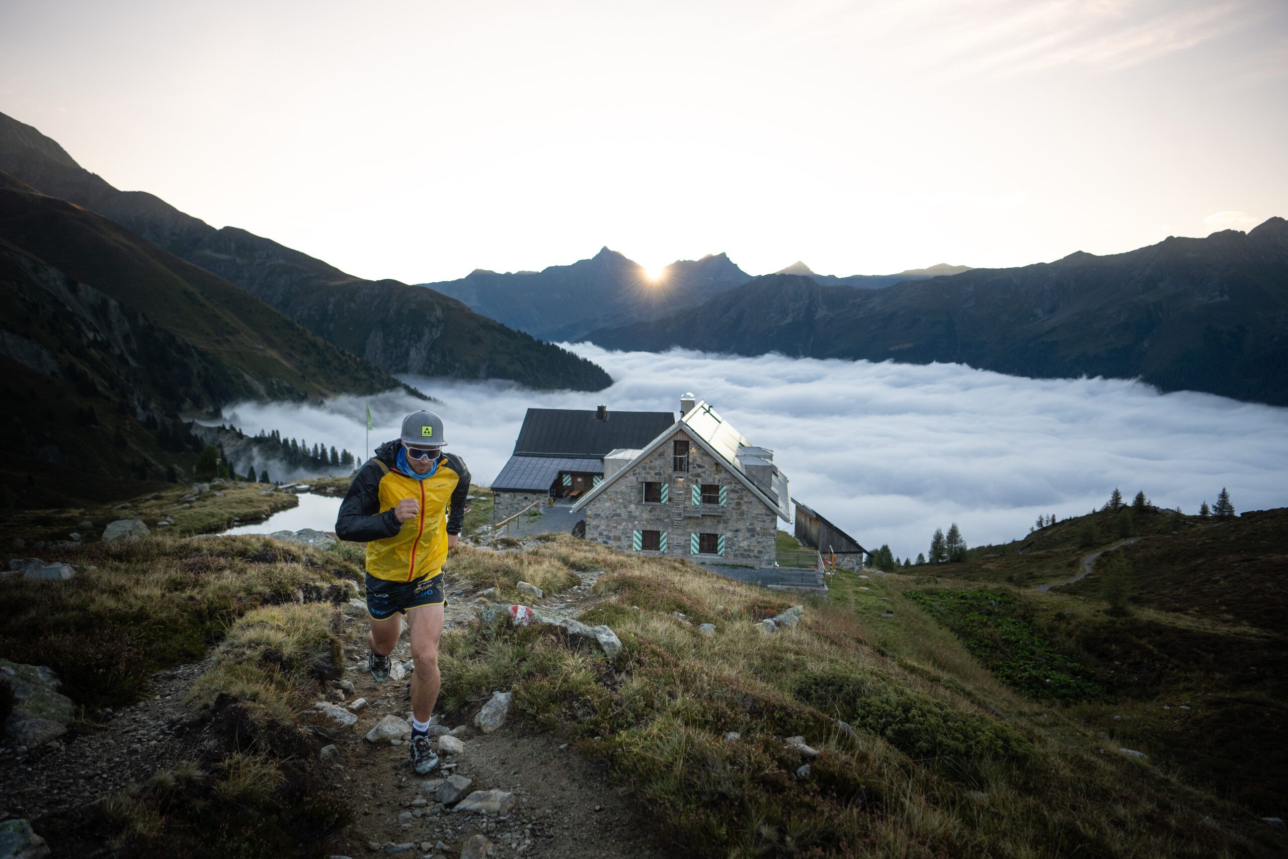 Trailrunner läuft einen Pfad hinter einer Berghütte hoch, Dahinter geht die Sonne über den Bergen und einer Nebeldecke im Tal auf